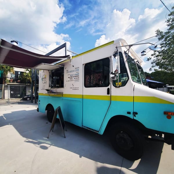 Latin Chicks food truck, white and teal with a yellow stripe, parked outdoors under a blue sky.