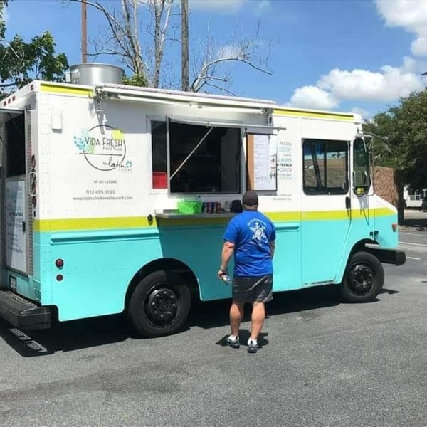 A man stands before the Latin Chicks "Vida Fresh" food truck, painted white, yellow, and teal.