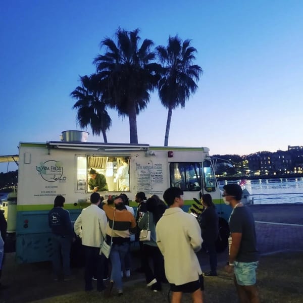 People gather around the Latin Chicks food truck at dusk, with palm trees and a river in the background.