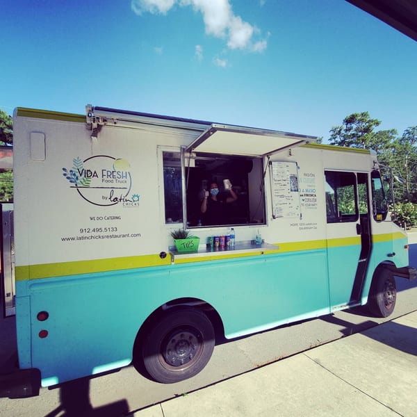 A Latin Chicks food truck, white with a teal and yellow stripe, under a blue sky.