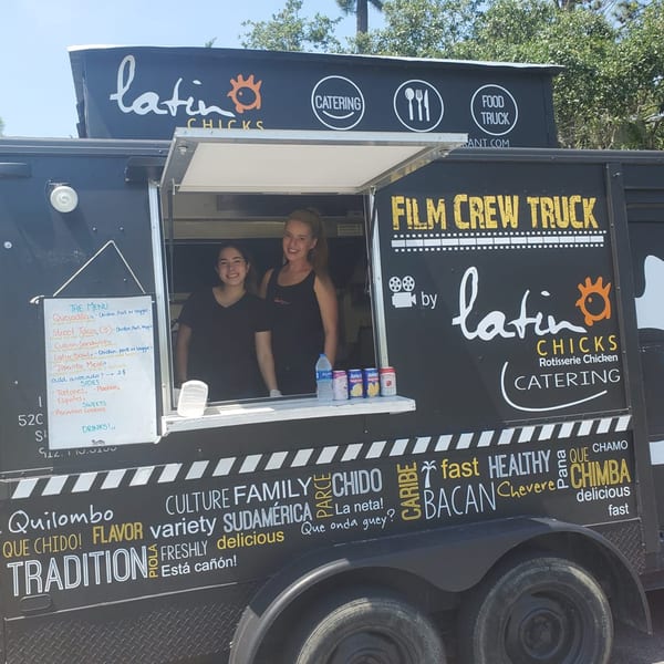 Two women smiling from inside the Latin Chicks food truck, with a menu and drinks visible.