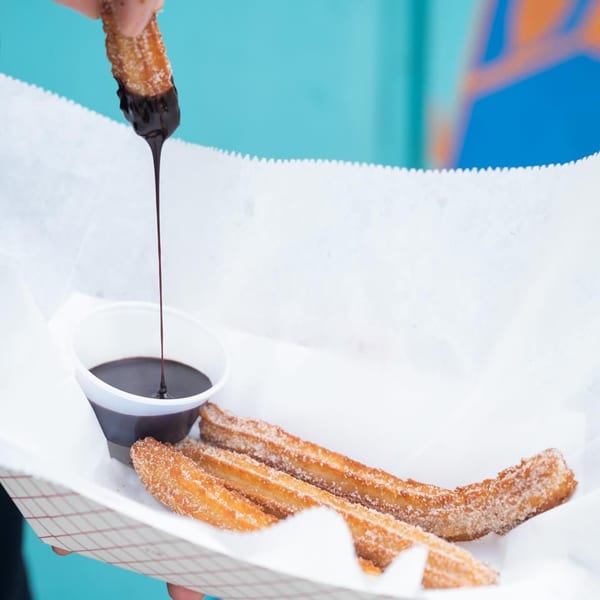 A hand dips a churro into a cup of chocolate sauce, with more churros in a paper tray.